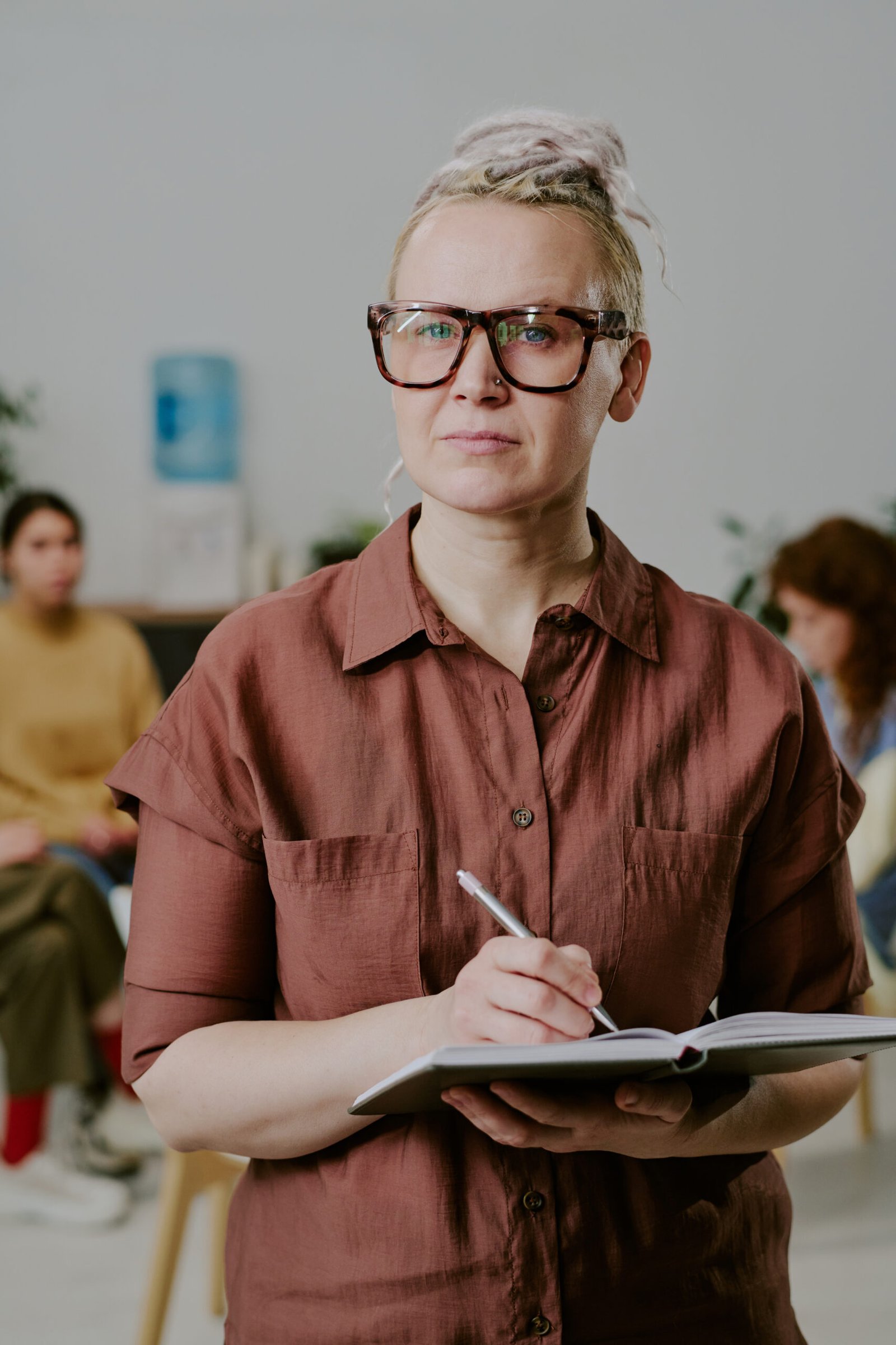 Portrait of confident woman holding notebook in office setting. Group of people sitting in background and engaging in discussion